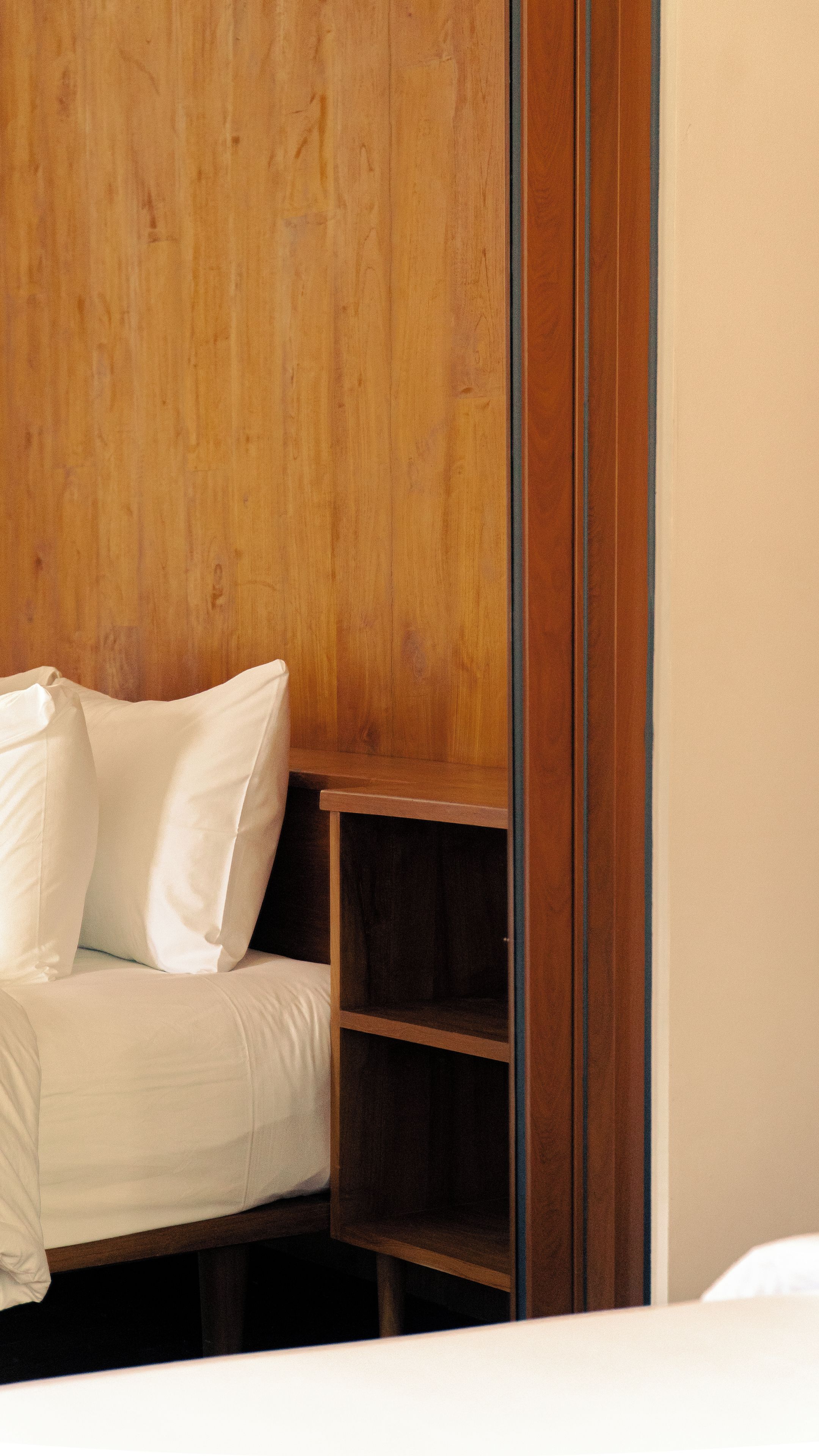 A bed with white bedding and pillows, a wooden headboard, and a small wooden shelf unit.