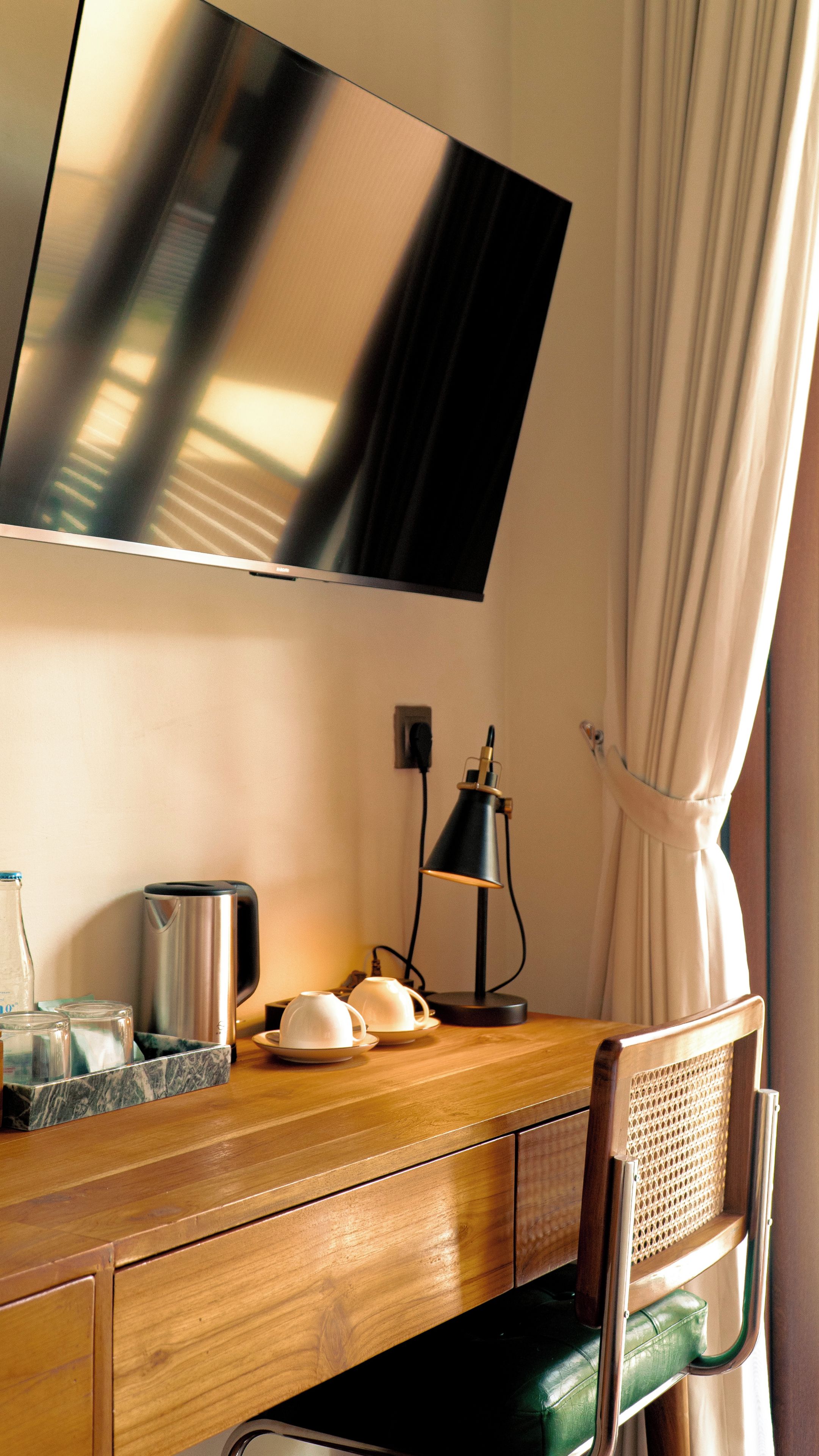 A hotel room with a wooden desk, a flat-screen TV mounted on the wall, a lamp, a kettle, and a chair with a green cushion.
