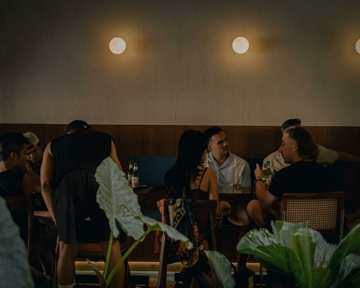 Eine Gruppe von Menschen sitzt bei gedämpftem Licht an einem Tisch in einem Raum mit schlichter Wand und zwei Deckenleuchten.