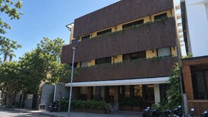A multi-story building with a modern design, featuring balconies with greenery, a covered entrance, and a street view with parked motorcycles.