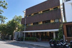 A multi-story building with a modern design, featuring balconies with greenery, a covered entrance, and a street view with parked motorcycles.