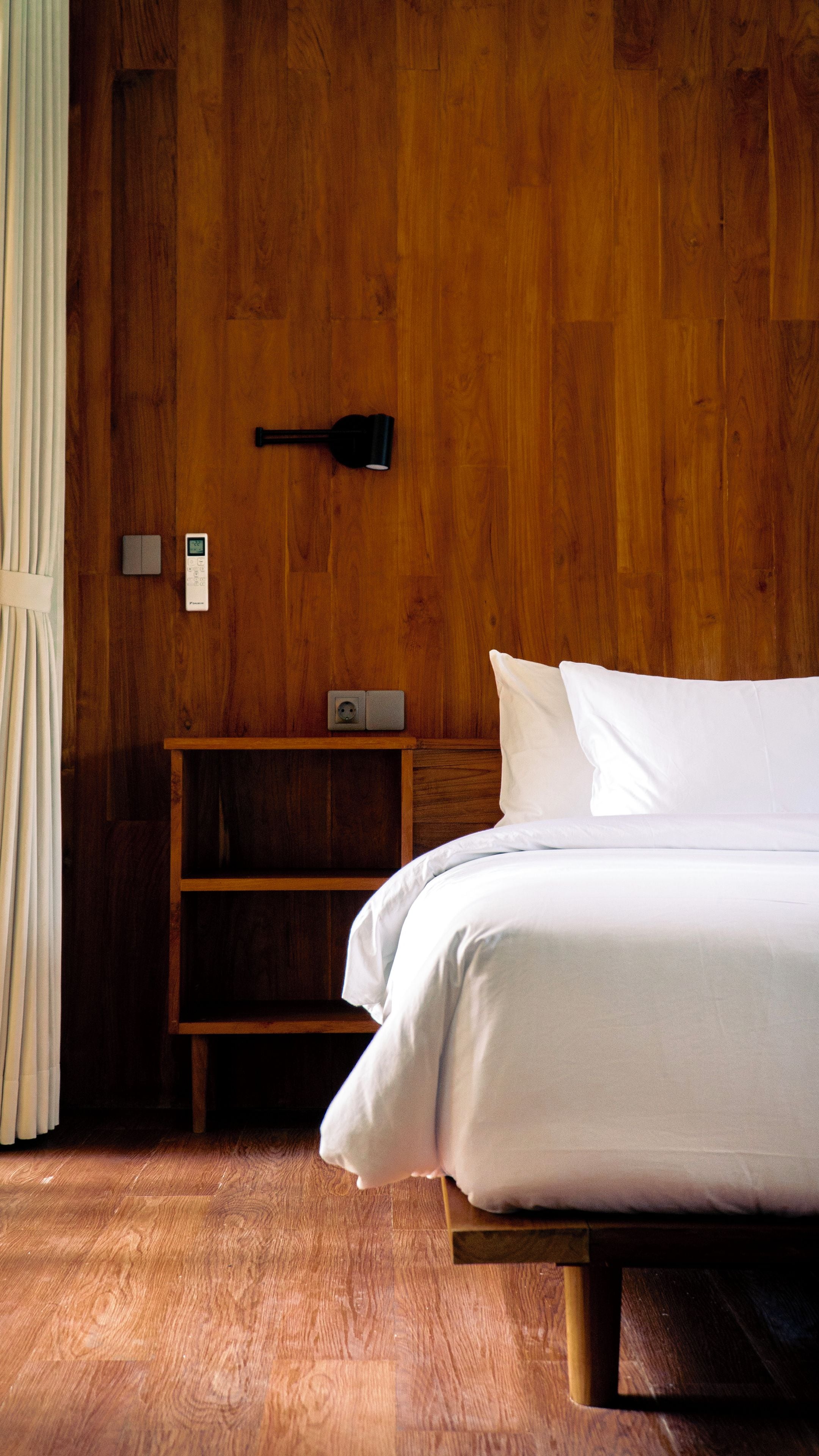 A bedroom with a wooden headboard, a bedside table with a lamp, a power outlet, and a bed with white bedding.