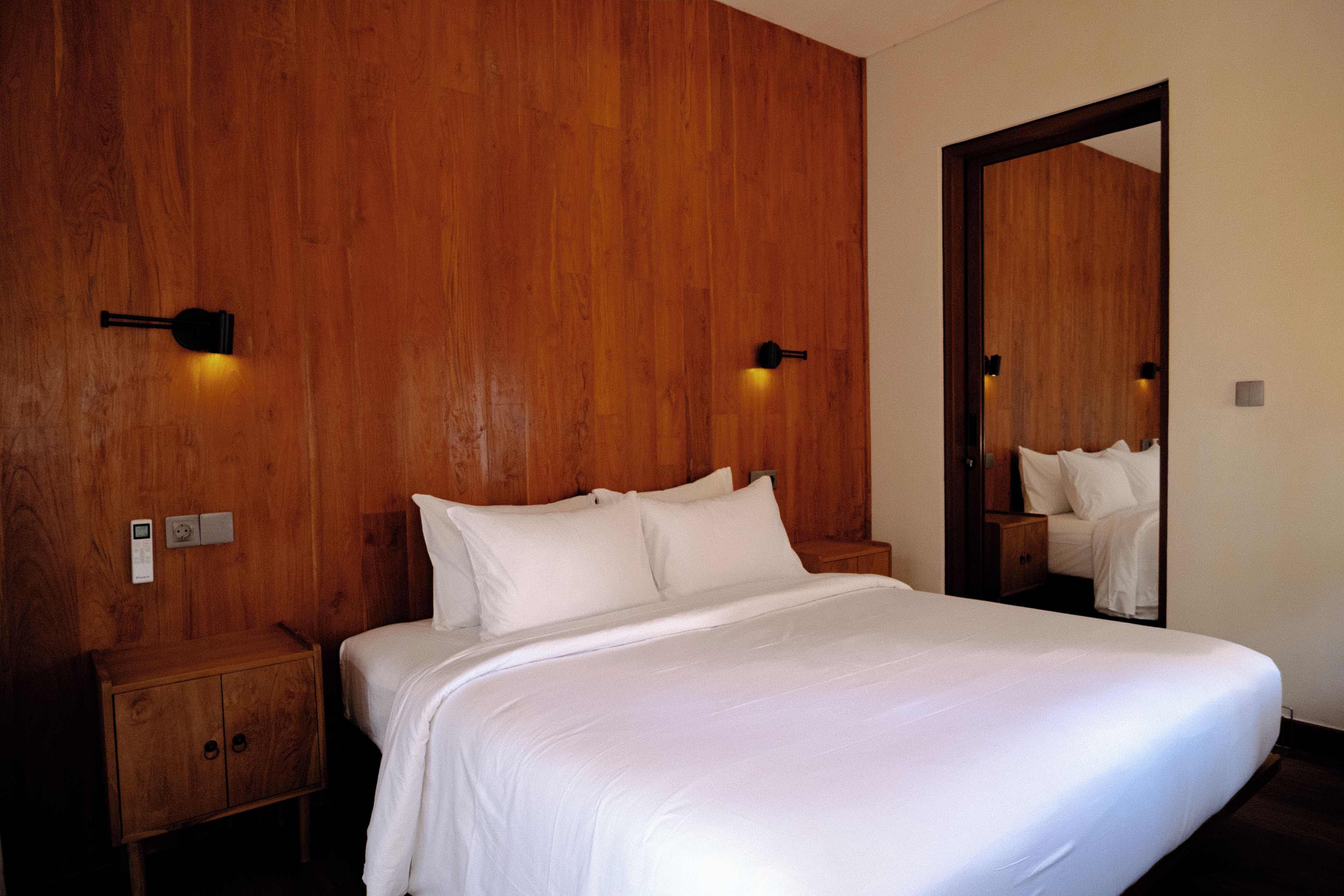 A hotel room with a wooden headboard, a bed with white linens, a nightstand, a bedside table, and a mirror on the wall.