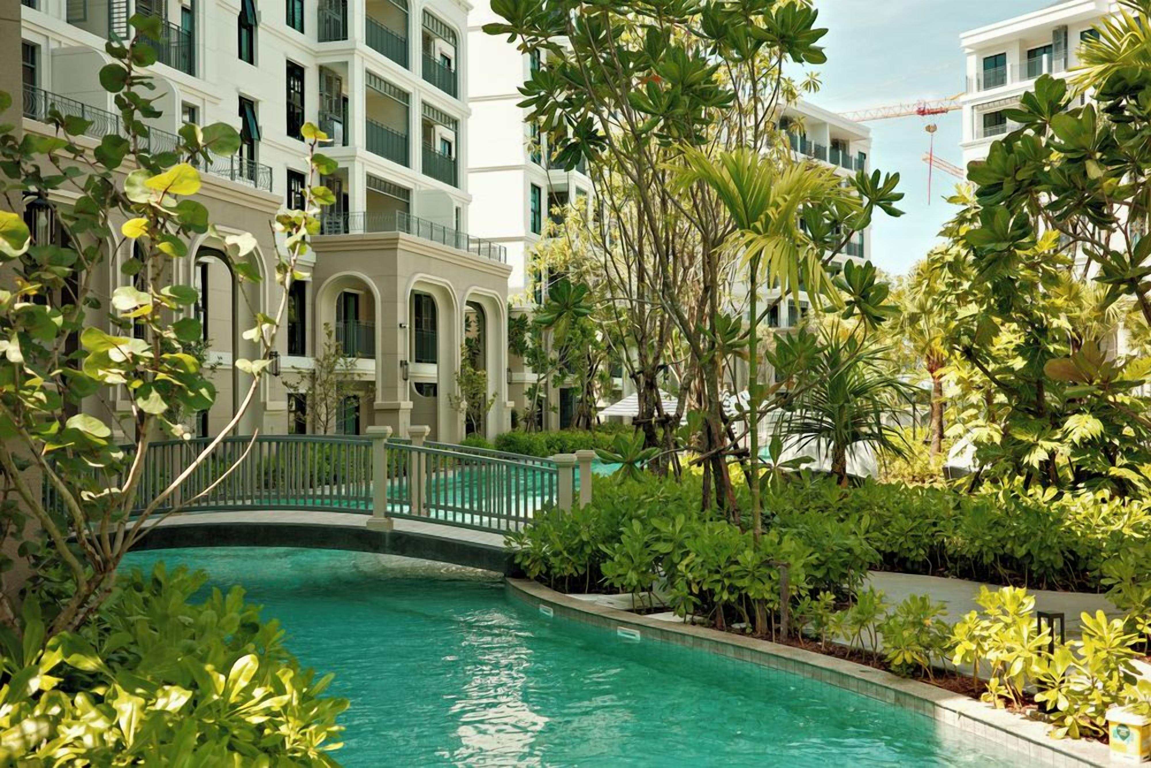 A pool surrounded by lush greenery and a white building with balconies.