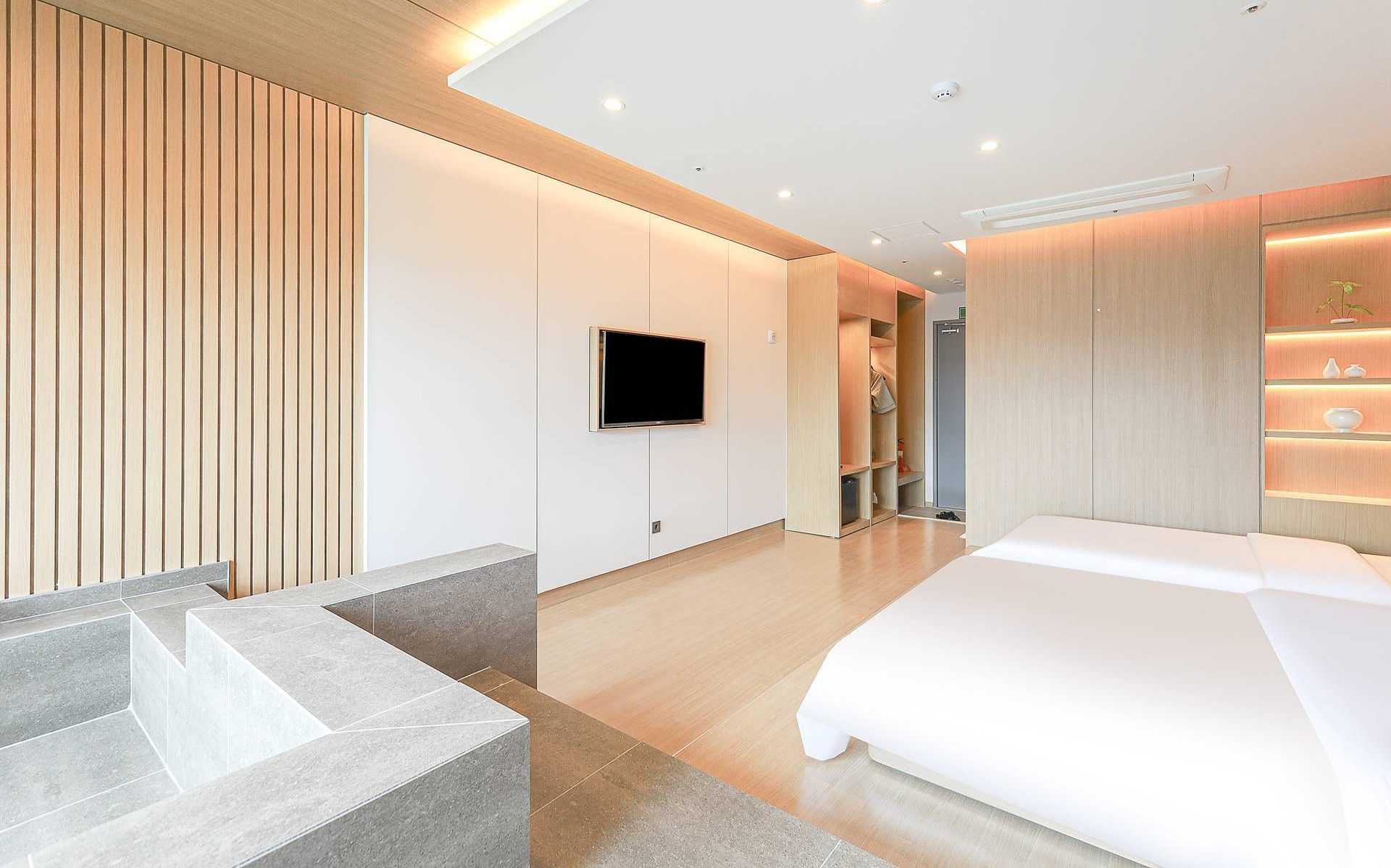 Una habitación de hotel moderna con cama, televisor montado en la pared, armario empotrado y una pequeña zona de estar con un banco.