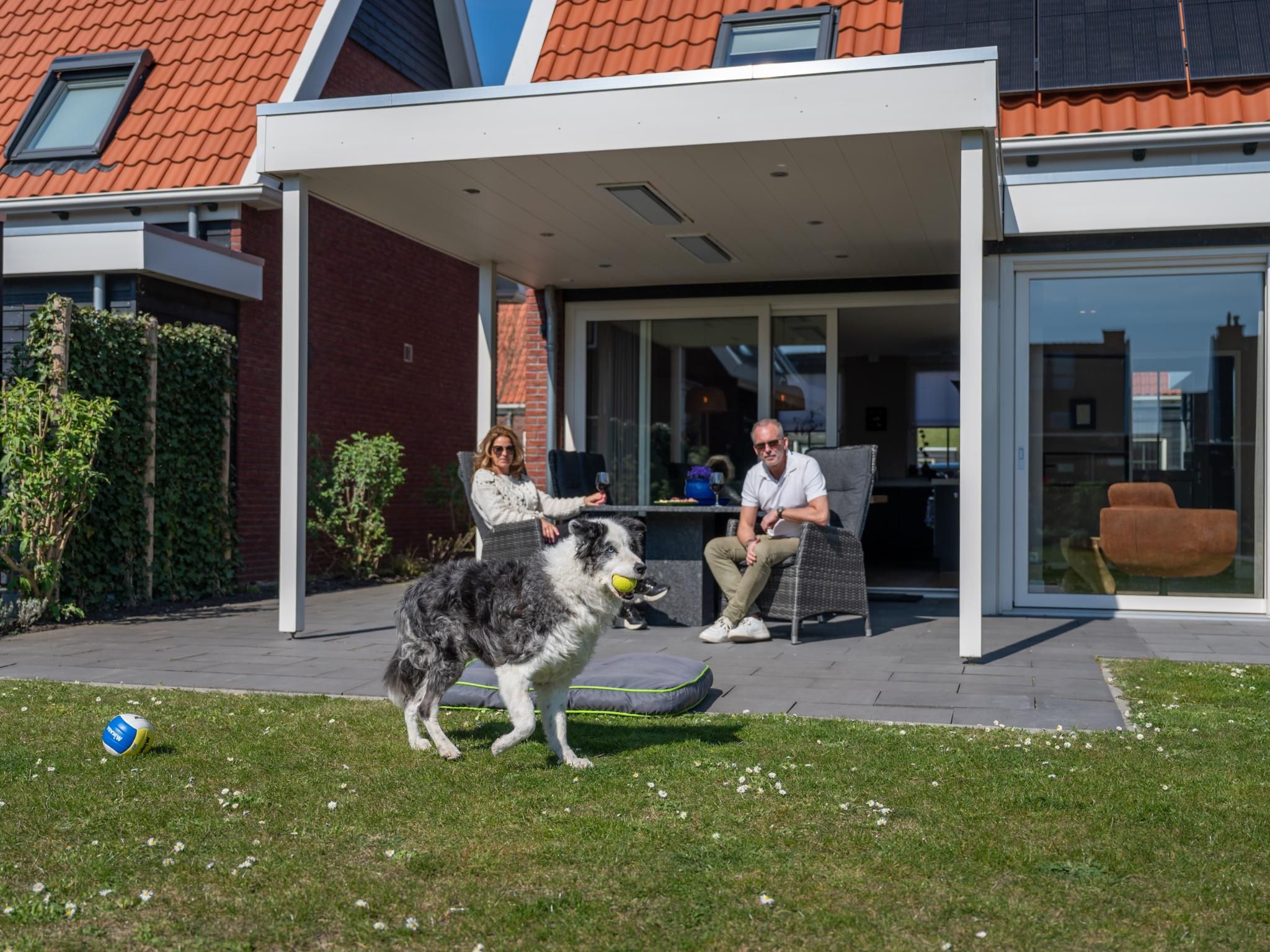 A modern house with a covered patio, a dog playing with a ball, and two people sitting on outdoor furniture.