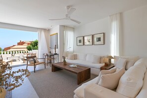 Ein geräumiges Wohnzimmer mit einer großen Couch, einem Holztisch und einem Deckenventilator. Durch die Fenster ist ein Balkon mit Gartenmöbeln zu sehen.
