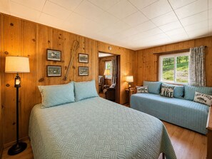 Une pièce aux murs en bois, avec un grand lit, deux petits lits, une lampe et des tableaux encadrés.