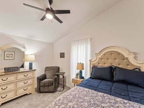 Ein Schlafzimmer mit einem Bett, einem Sessel, einer Kommode, einem Nachttisch, einem Deckenventilator und einem Fenster mit Vorhängen.