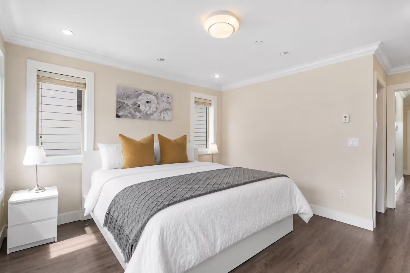 Une chambre à coucher comprenant un lit, deux tables de chevet, une fenêtre avec des stores, un luminaire au plafond et une œuvre d’art accrochée au mur.