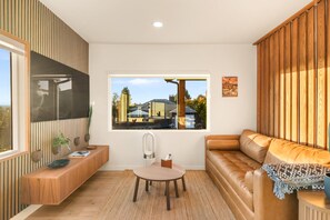 A modern living room with a large window, a wooden wall panel, a sofa, a small round table, and a TV mounted on the wall. - Private Softub, Walk to Junction - 2BR + Den, King Bed (Seattle)