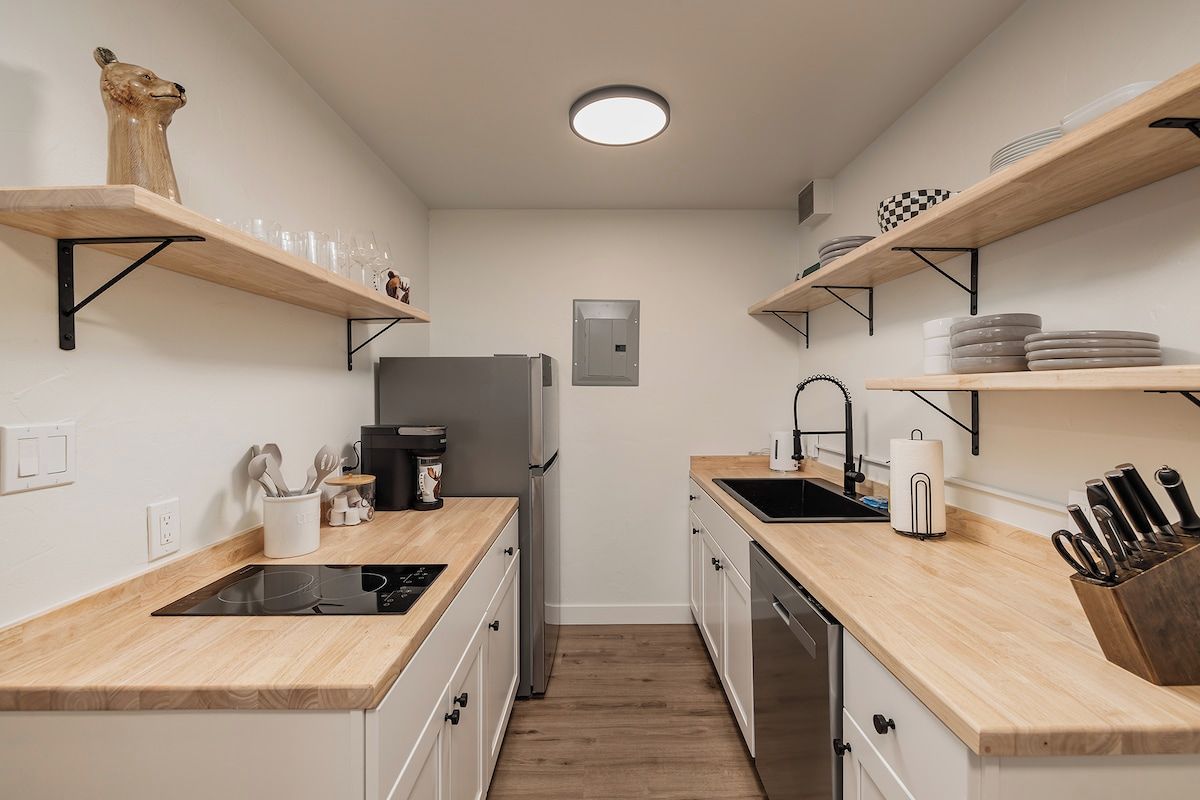 Una cocina compacta con encimeras de madera, horno integrado, refrigerador y estantes con vajilla y utensilios.