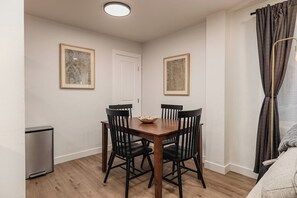 A dining area with a wooden table and four chairs, a small trash bin, and two framed pictures on the walls. - Downtown Anchorage Studio - King Bed -Sleeps 4 (Anchorage)