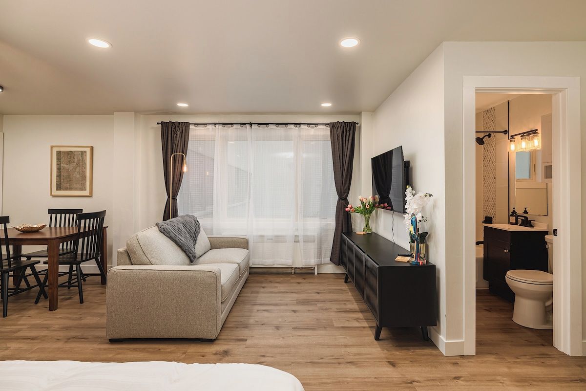 Ein Hotelzimmer mit einer Couch, einem Esstisch mit Stühlen, einem Fernseher, einem Konsolentisch mit einer Vase und einem durch eine offene Tür einsehbaren Badezimmer.
