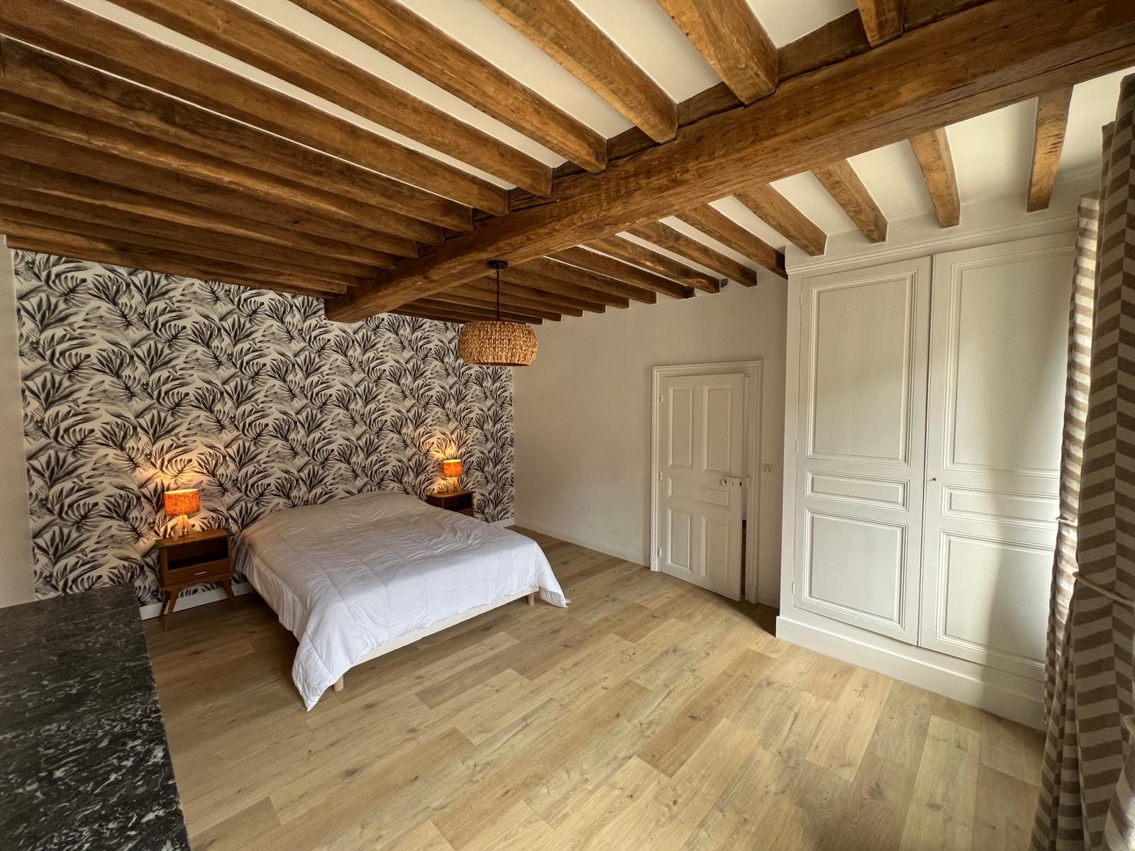 Un dormitorio con cama, techo de madera, papel tapiz estampado, puerta blanca y cortina a rayas.