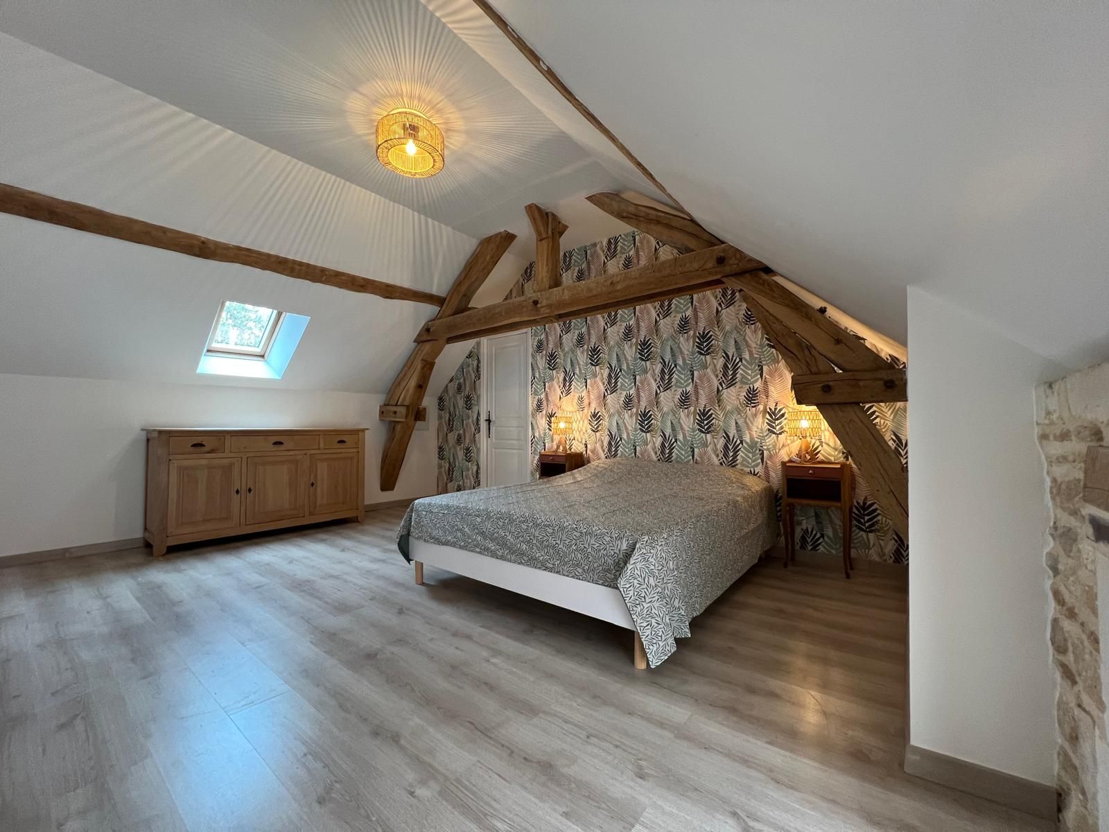 Quarto com teto inclinado, vigas de madeira, cama com roupa de cama estampada, guarda-roupa e uma pequena janela.