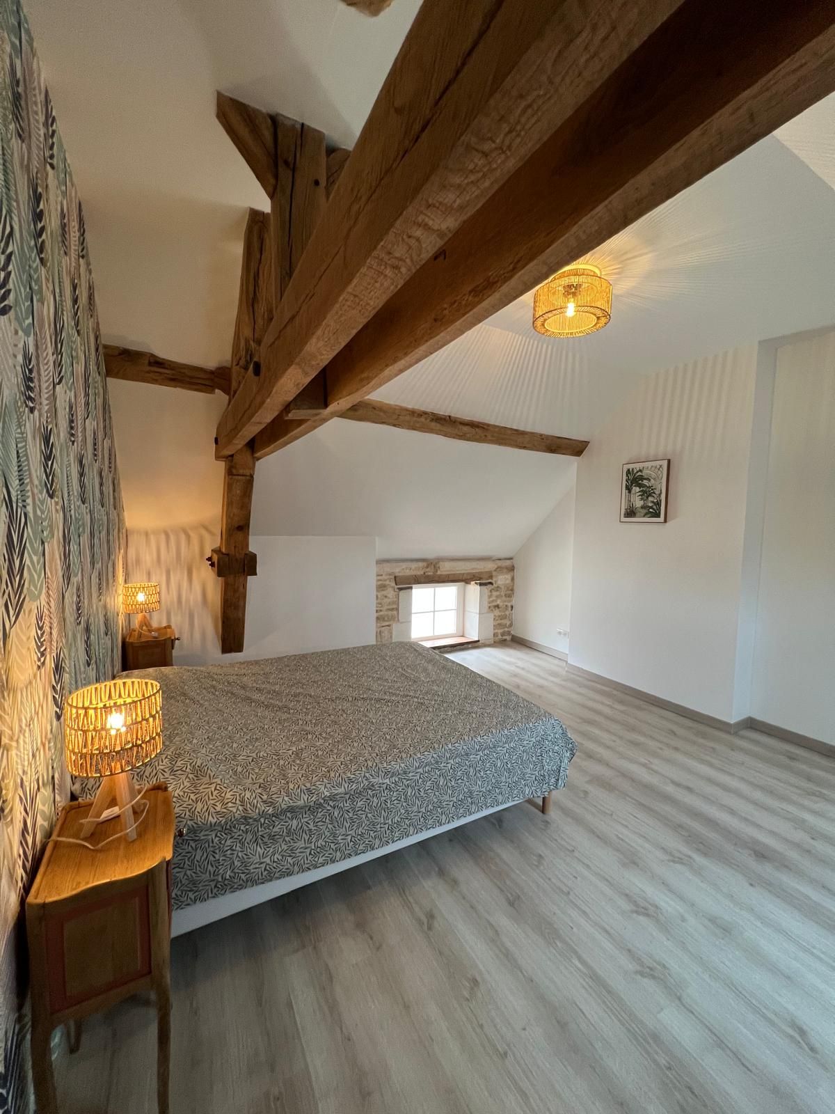 Un dormitorio amplio en el ático con vigas de madera a la vista, una colcha estampada y un ventanal pequeño.