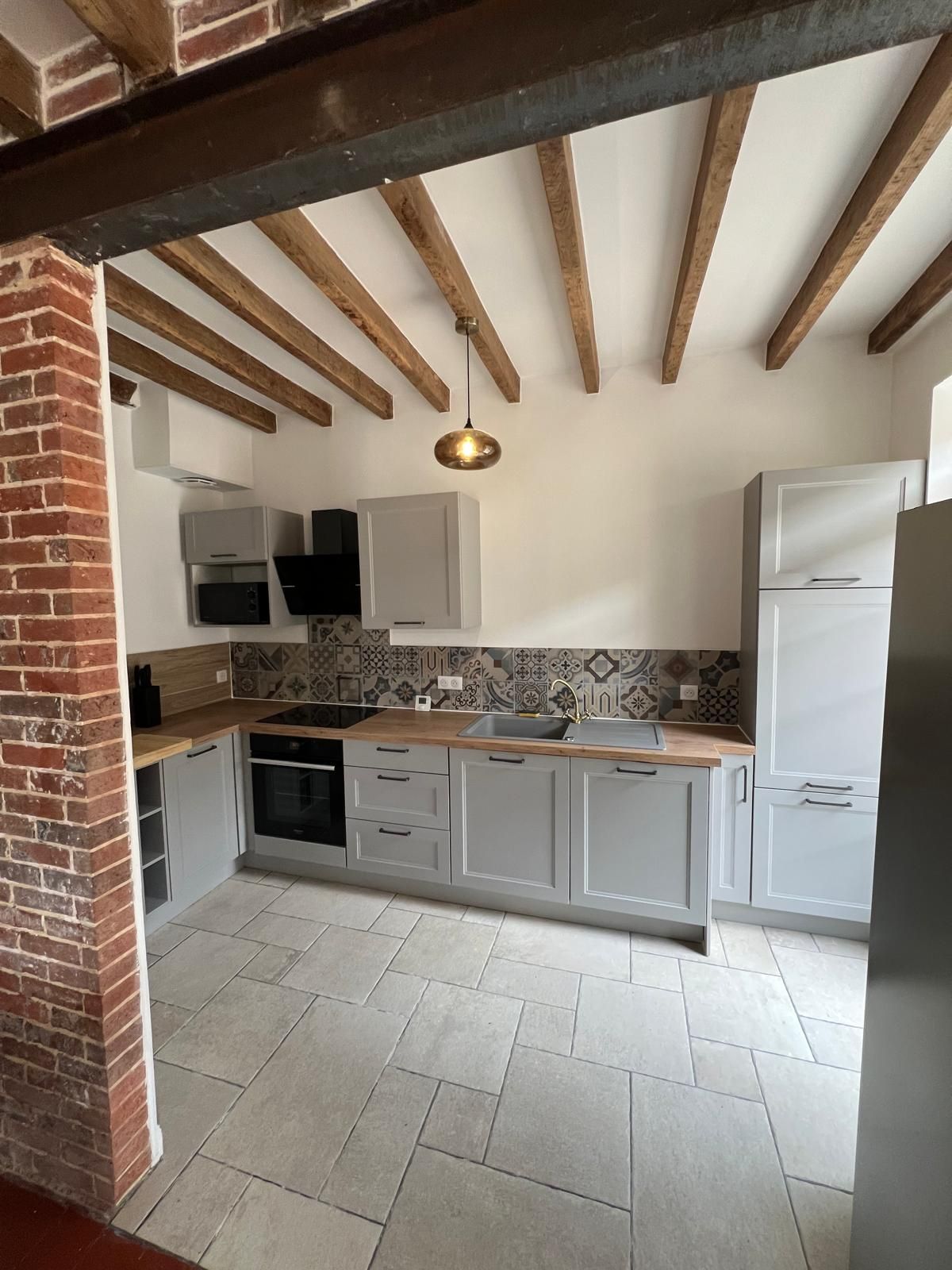 A kitchen with a brick column, wooden beams, a hanging light fixture, and a tiled backsplash.