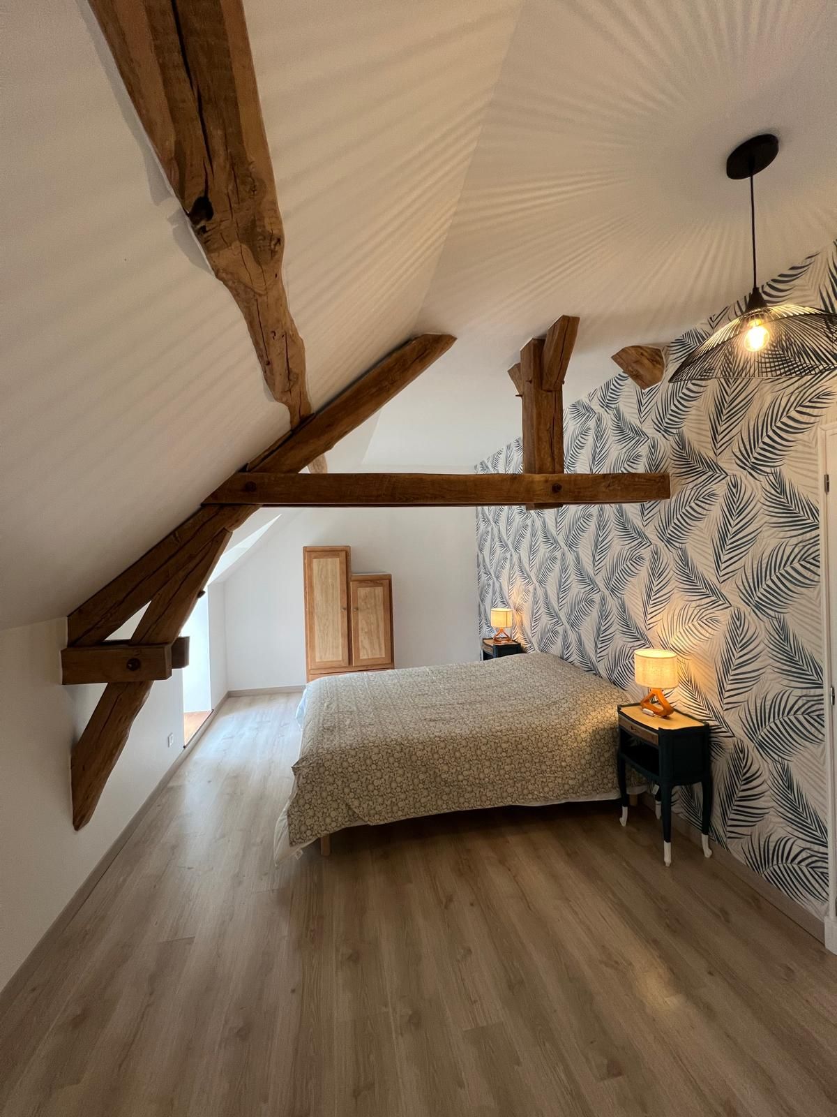 A bedroom with a sloped ceiling, wooden beams, a bed, a nightstand, and a patterned wall.