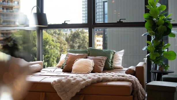 Een moderne slaapkamer met een groot raam, een bed met veel kussens, een nachtkastje en een potplant.