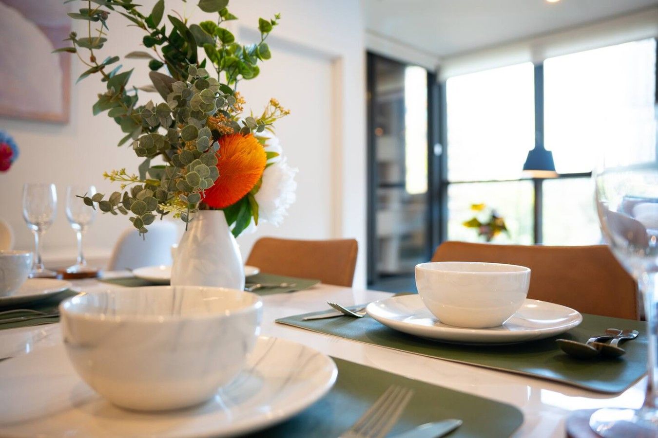 A dining table set with white bowls, a vase with greenery and an orange flower, and a window with natural light.