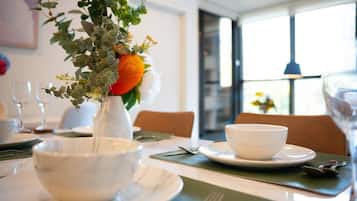 A dining table set with white bowls, a vase with greenery and an orange flower, and a window with natural light.