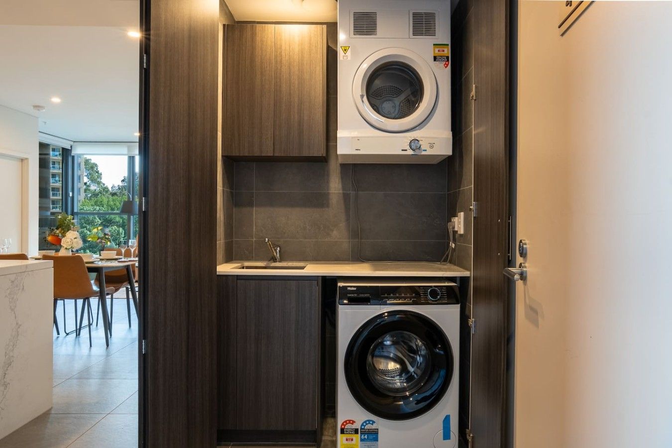 Een praktische wasruimte met een wasmachine en droger, een klein wastafeltje, en uitzicht op een eethoek met een tafel en stoelen.
