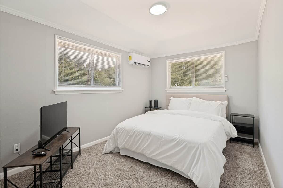 Une chambre à coucher avec un lit, un téléviseur posé sur un meuble et deux fenêtres équipées de stores.