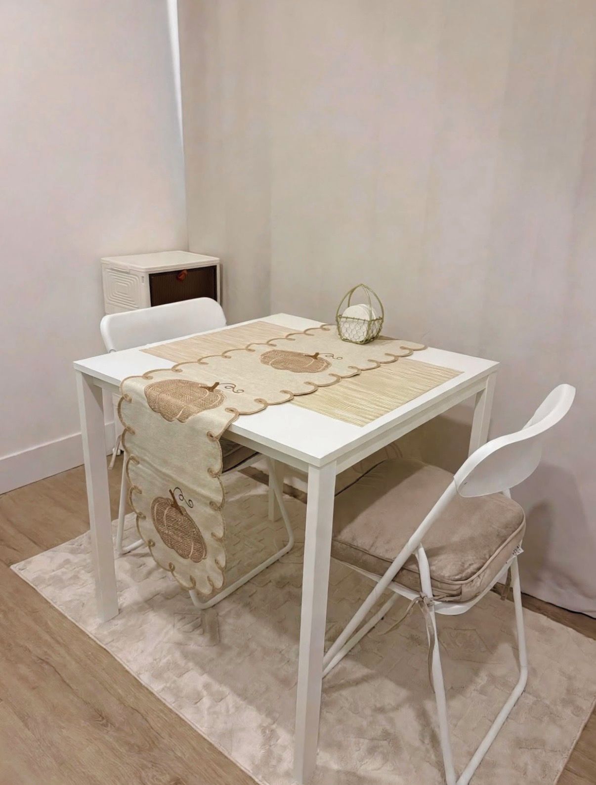 A white table with a woven placemat and a decorative cushion, accompanied by a white chair with a cushioned seat.