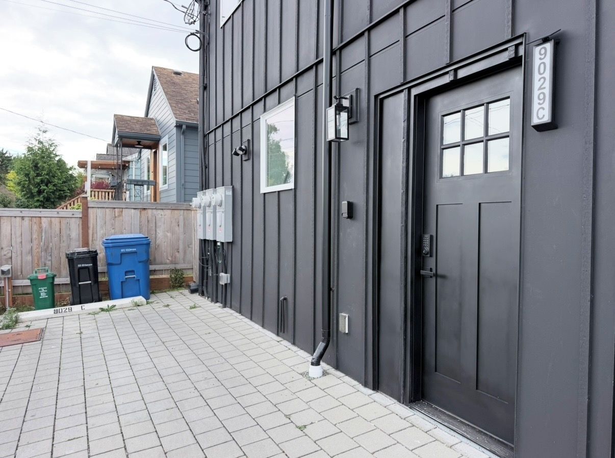 A building with a dark exterior, a door with a number plate, and a paved patio area with trash bins.