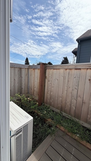 En træterrasse med en metal klimaanlæg, et træhegn og en klar himmel med skyer.