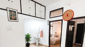 Une pièce spacieuse avec un plancher en bois, un mur blanc orné d’un tableau encadré, une plante en pot et un lit avec une table de chevet et une lampe.