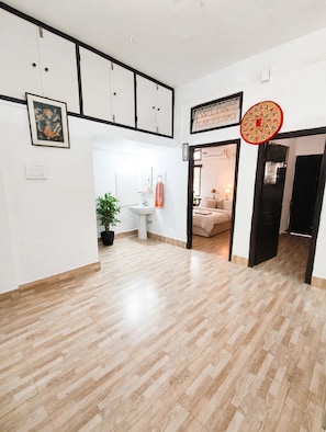 A spacious room with a wooden floor, a white wall with a framed picture, a potted plant, and a bed with a bedside table and lamp. - Vandara Oasis | King Size Bed AC Room (Guwahati)