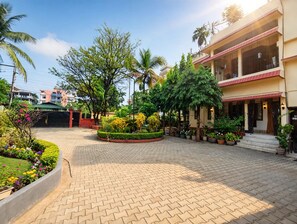 A courtyard with a paved walkway, surrounded by greenery and palm trees, leading to a building with a red roof and multiple balconies. - Vandara Oasis | King Size Bed AC Room (Guwahati)