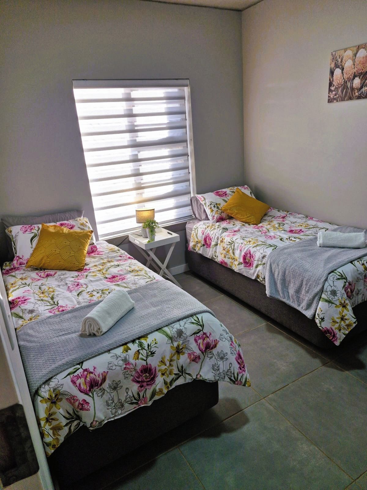 A room with two beds, each with floral bedding and a single pillow. There is a small table with a lamp and a plant.