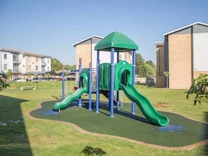 A playground with slides and climbing structures, surrounded by a grassy area and residential buildings.
