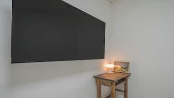 Una habitación con una amplia pantalla de proyección negra en la pared, una pequeña mesa de madera con una lámpara y un cuadro enmarcado, además de una pared blanca.
