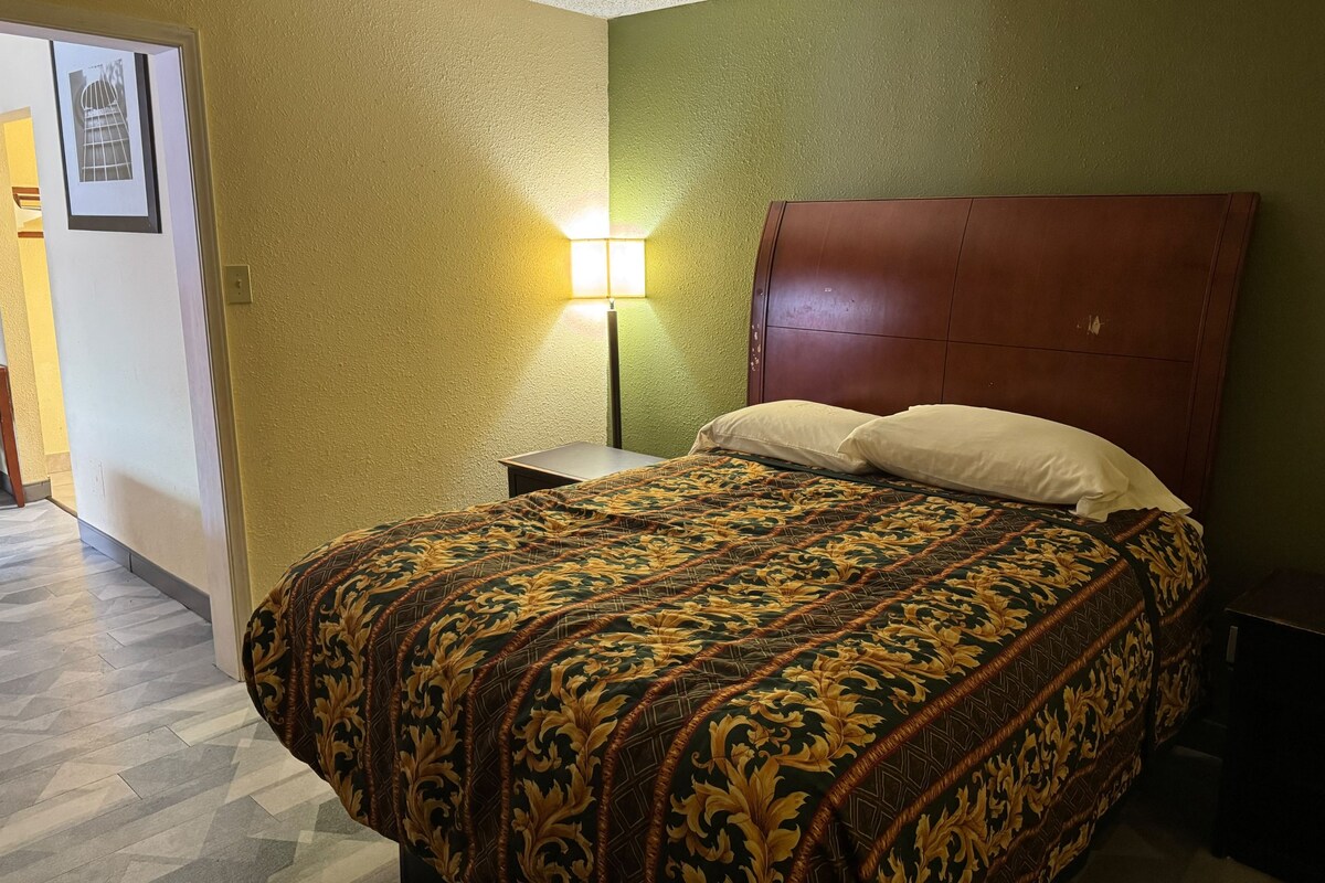 A hotel room with a bed, a nightstand, a lamp, and a door.