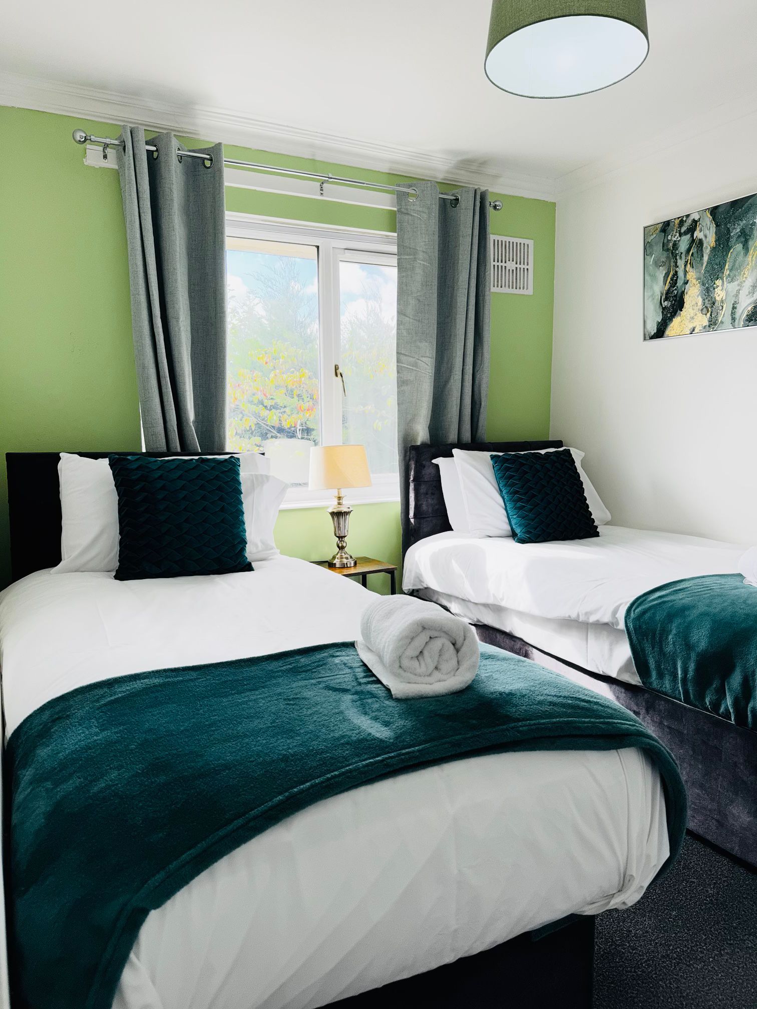 Ein Hotelzimmer mit zwei Betten, einem Fenster mit Vorhängen, einer Nachttischlampe und einem Bild an der Wand.