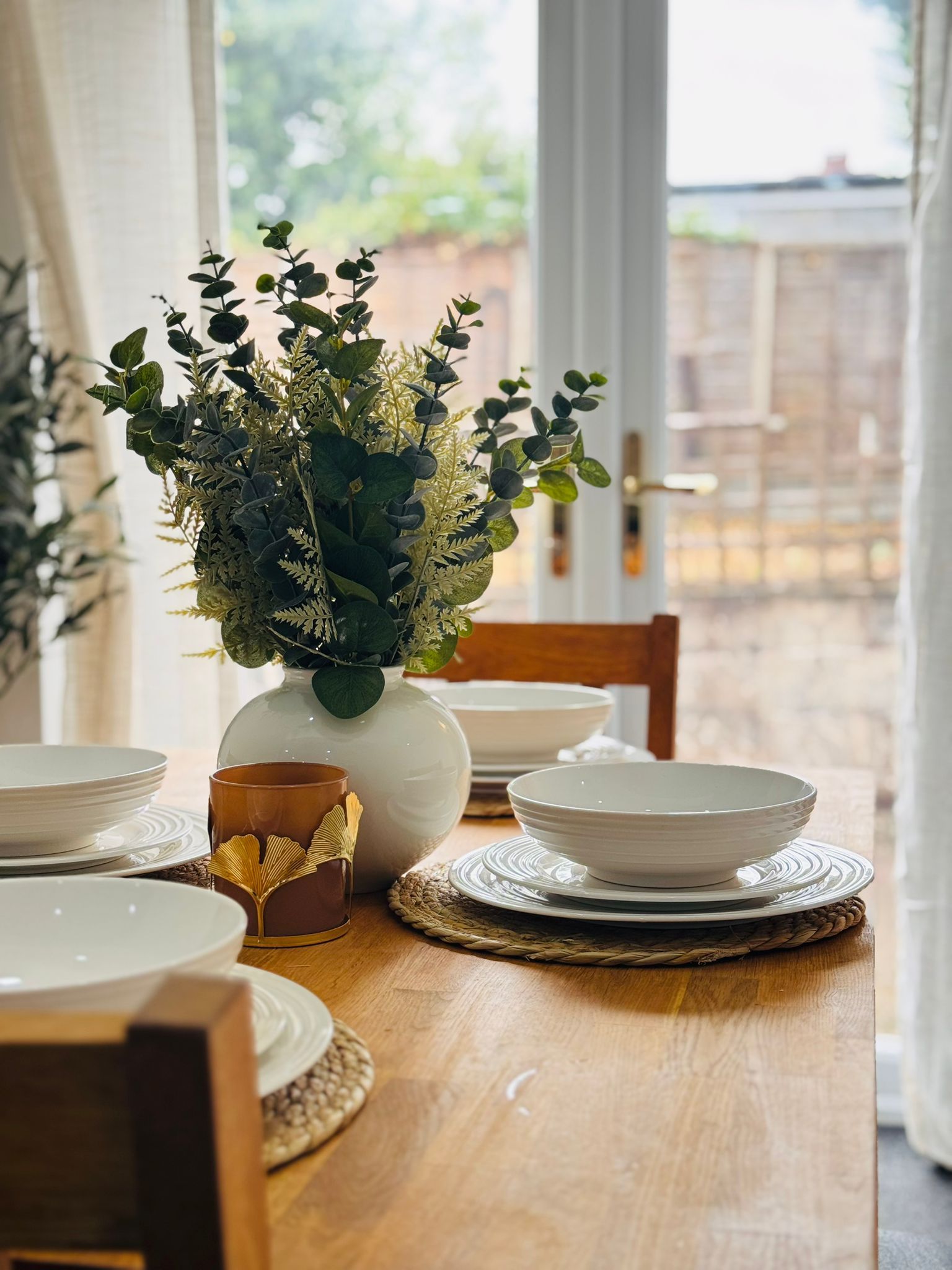 A dining table with a white vase containing green foliage, a brown mug, and white plates and bowls.