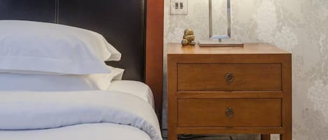 A neatly made bed with white linens, a wooden nightstand with a lamp, and a small shelf with books.