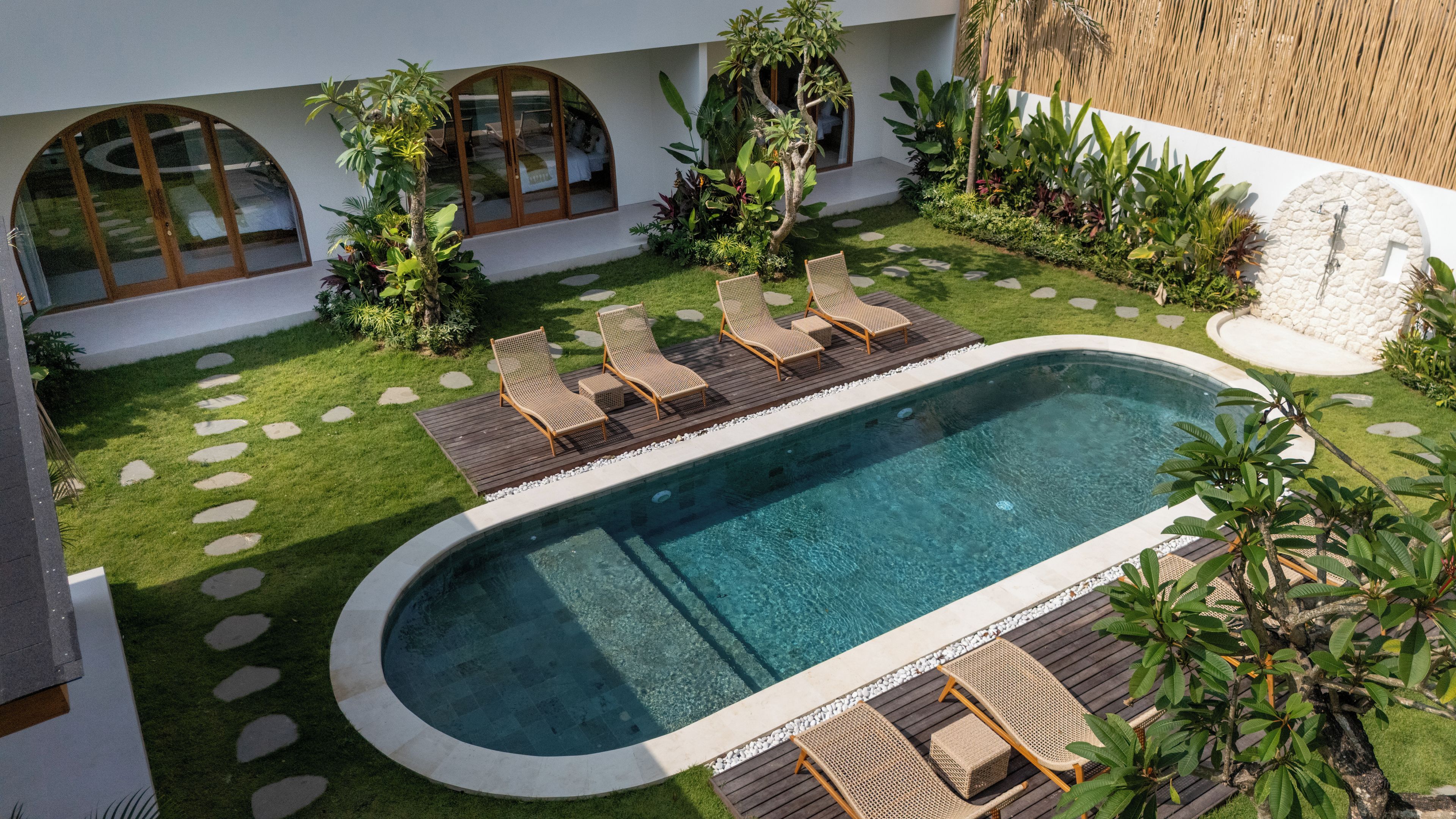 Una piscina con sillas de descanso, rodeada de césped cuidado y plantas tropicales.