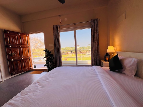 A hotel room with a large bed, a nightstand with a lamp, a window with curtains, and a view of the outside.