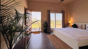 A hotel room with a bed, a ceiling fan, a large window with curtains, and a potted plant. - Habba Dates Village (CHIKKABALLAPURA)