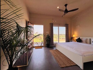 Quarto de hotel com cama, ventilador de teto, janela grande com cortinas e planta em vaso.