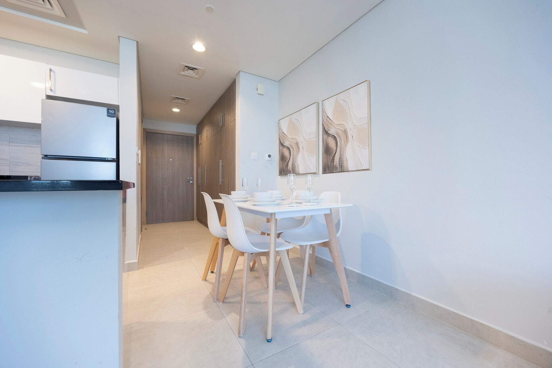 Una cocina moderna con un refrigerador empotrado, un comedor con una mesa y sillas blancas, y un pasillo que lleva a una puerta.