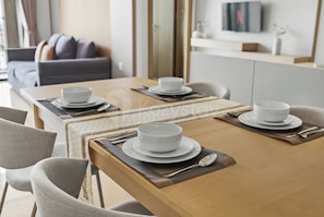 A dining table set with white plates and bowls, accompanied by cutlery, in a modern interior with a sofa and a TV in the background. - Chiro by Kozystay - Tanjung Barat (Jakarta)