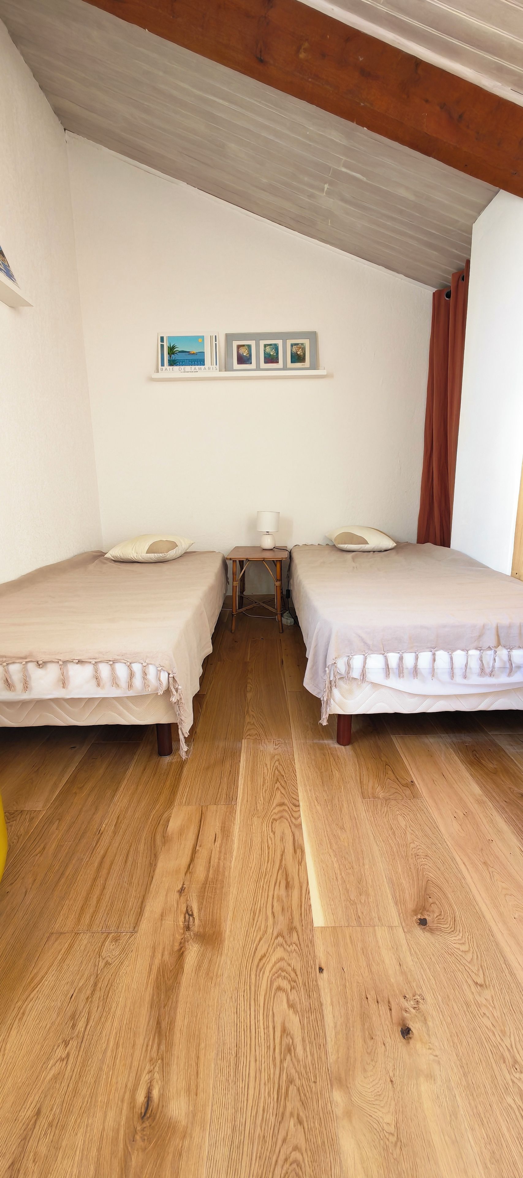 Une chambre avec deux lits, une petite table et une étagère fixée au mur, ornée de cadres avec des photos.