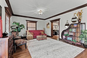 A living room with a red sofa, patterned ottomans, a large plant, and a wooden bookshelf with books and decorative items. - Historic Bruce Goff Home with Pool, Hot tub & Sauna near Utica Square (Tulsa)