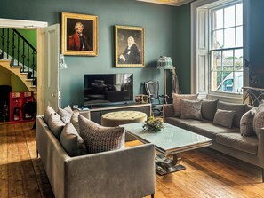 A living room with a sofa, ottoman, and a coffee table. There is a television, a lamp, and two framed pictures on the wall. A staircase is visible in the background. - 4 bedroom accommodation in Montrose (Montrose)
